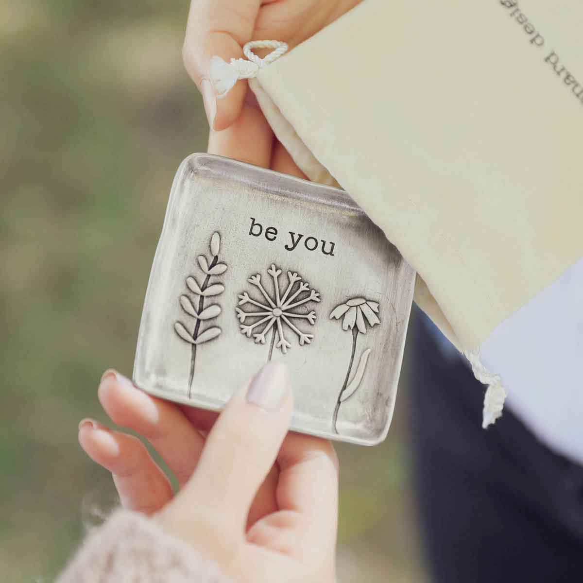 hands taking the be you pewter keepsake dish out from a lisa leonard bag