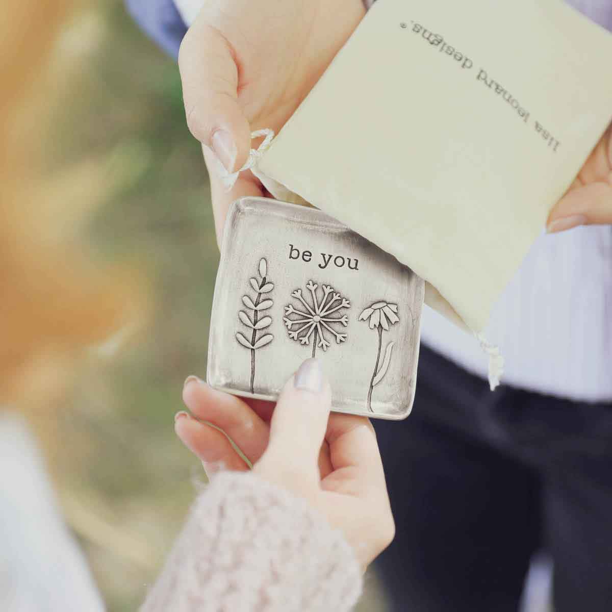 hands taking the be you pewter keepsake dish out from a lisa leonard bag