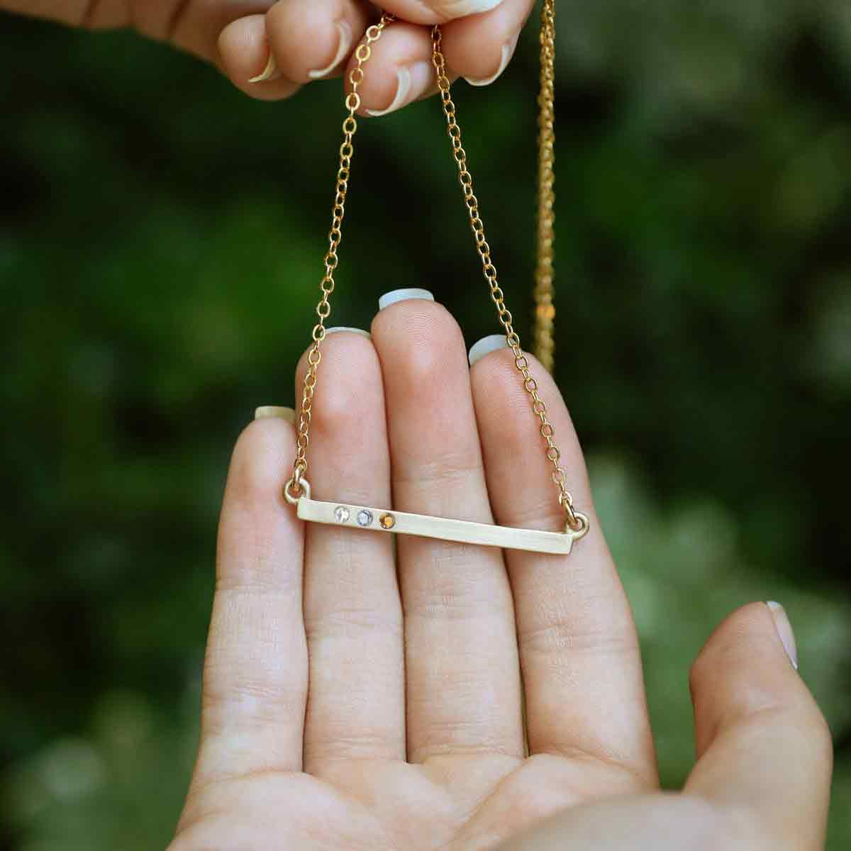 hand holding14k yellow gold cross bar birthstone necklace strung on a gold-filled link chain customizable with up to 7 genuine 2mm birthstones